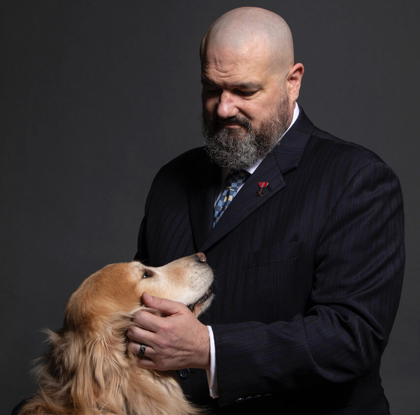 Man in a dark pinstripe suit gently holds a golden retriever’s snout, looking down affectionately; dog wears a service dog vest.