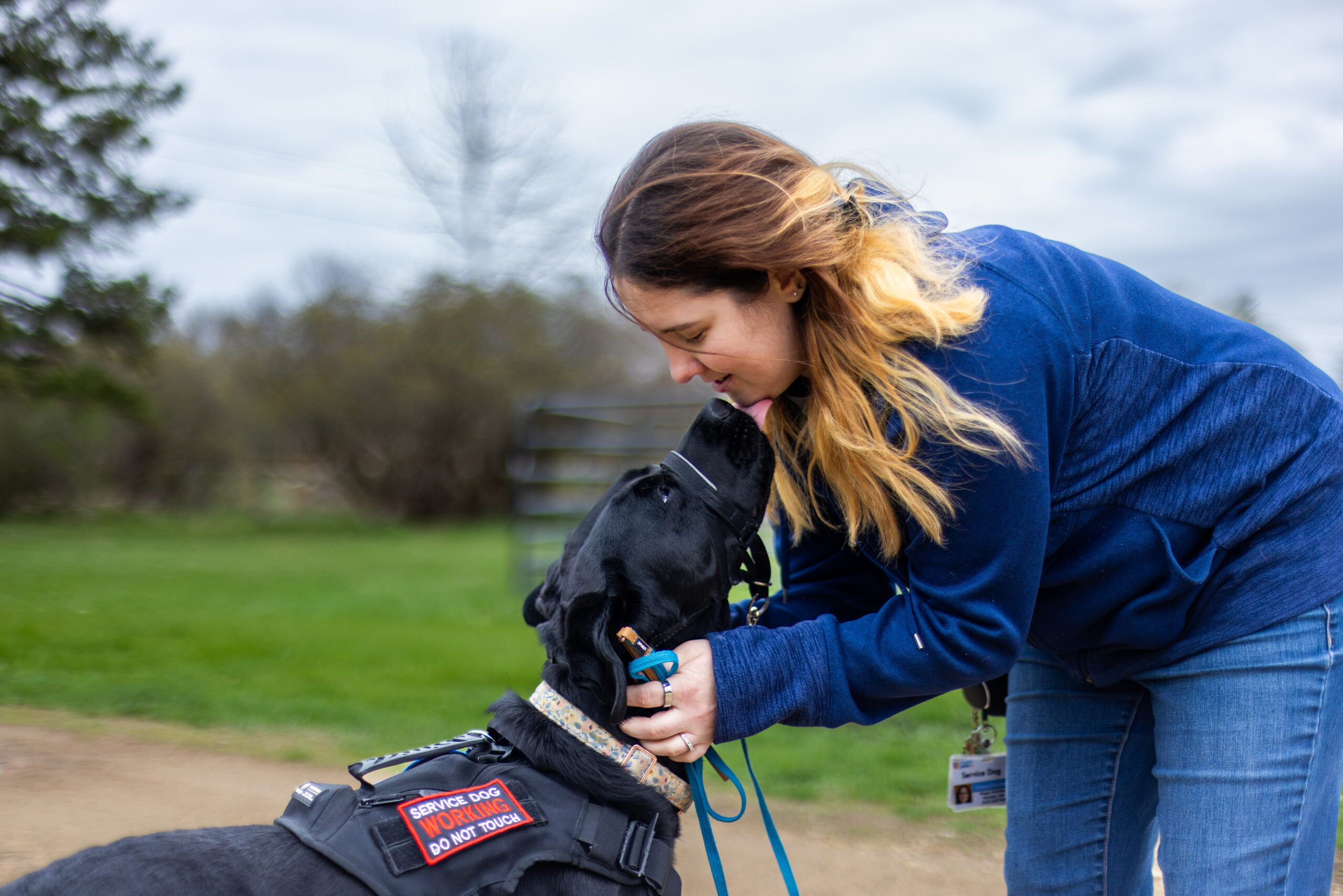 Megan Jesse-7 - Puppy Jake Foundation Person in a blue jacket leans down to hug a black service dog wearing a vest that reads 'SERVICE DOG WORKING DO NOT TOUCH' outdoors.