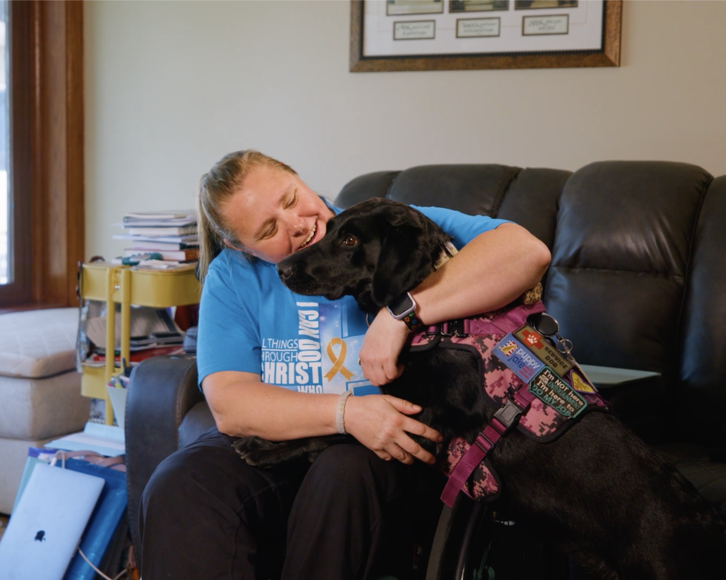 Person in a blue shirt hugging a large black dog on a brown leather couch in a living room.