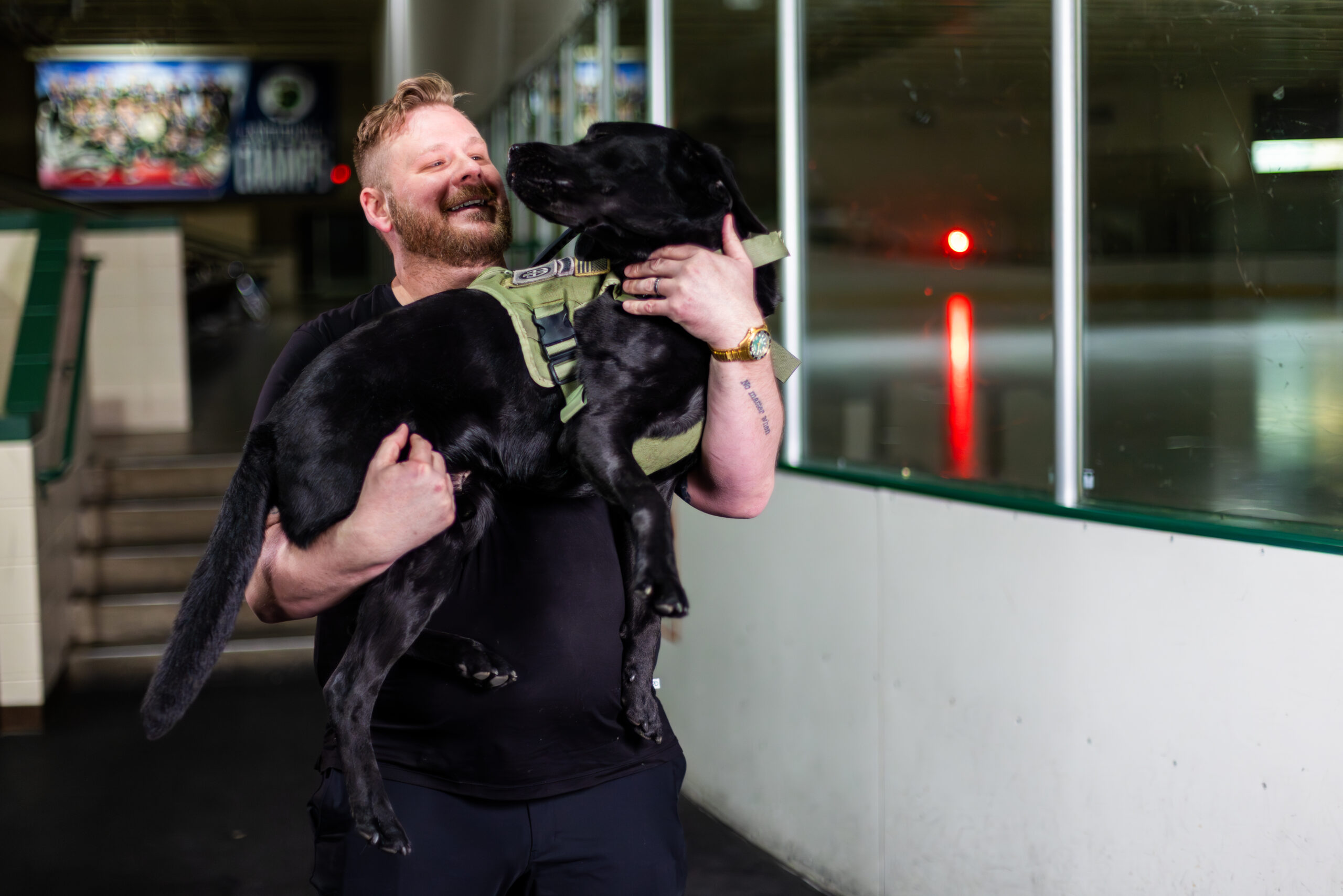 Sean Chips-3 - Puppy Jake Foundation Man in a black shirt smiles while lifting a large black dog in a green harness indoors, near glass doors and tiled walls.