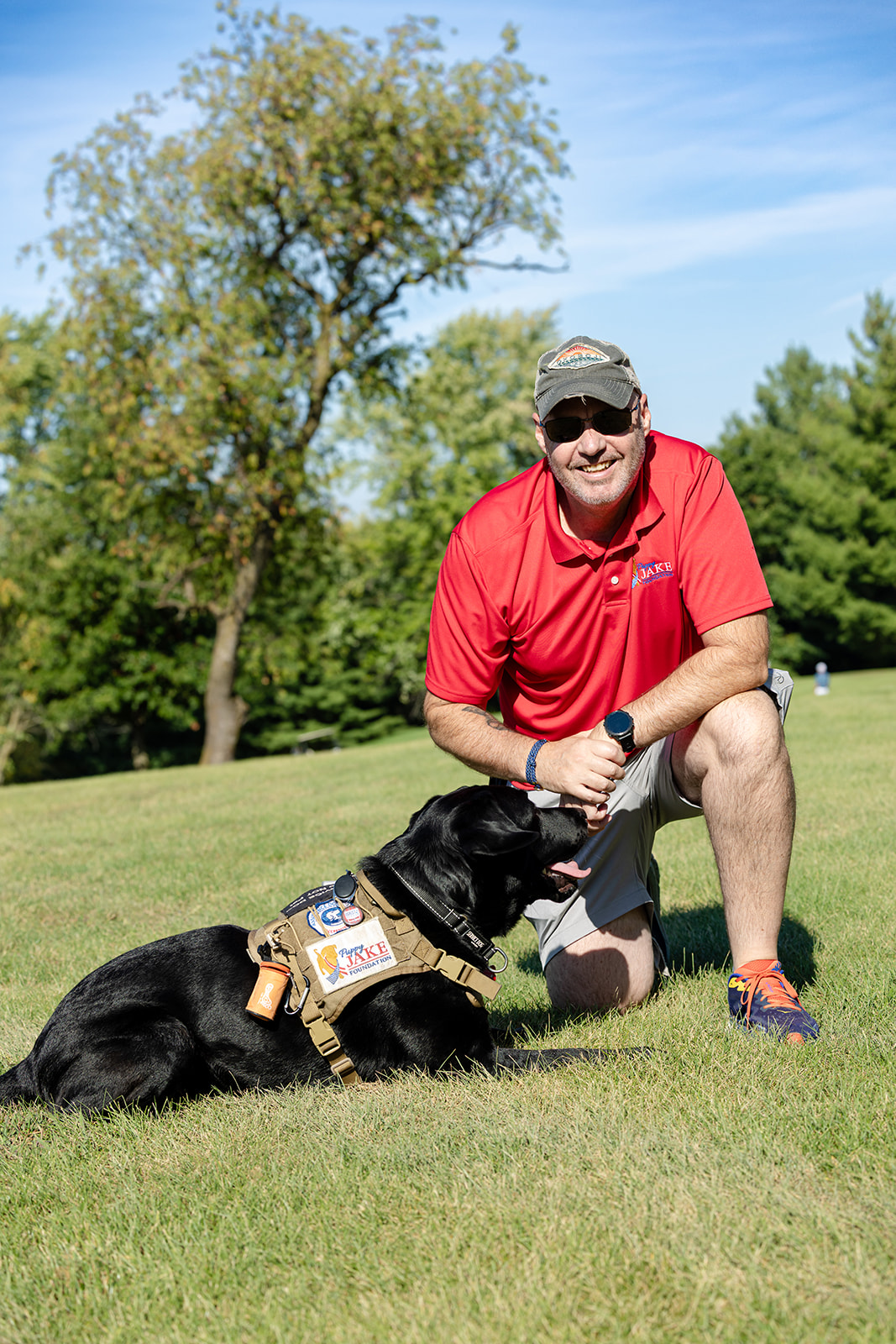 Puppy Jake 5th Annual Golf - Puppy Jake Foundation - Puppy Jake Foundation Man in a red polo kneeling on a grassy field, gently holding the paw of a black dog wearing a service/working vest with patches in a sunny park.