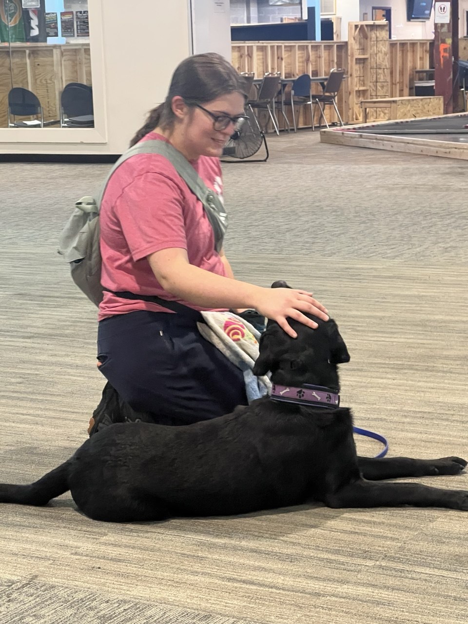 Woman in a pink shirt kneels and pets a large black dog on a beige carpet indoors.