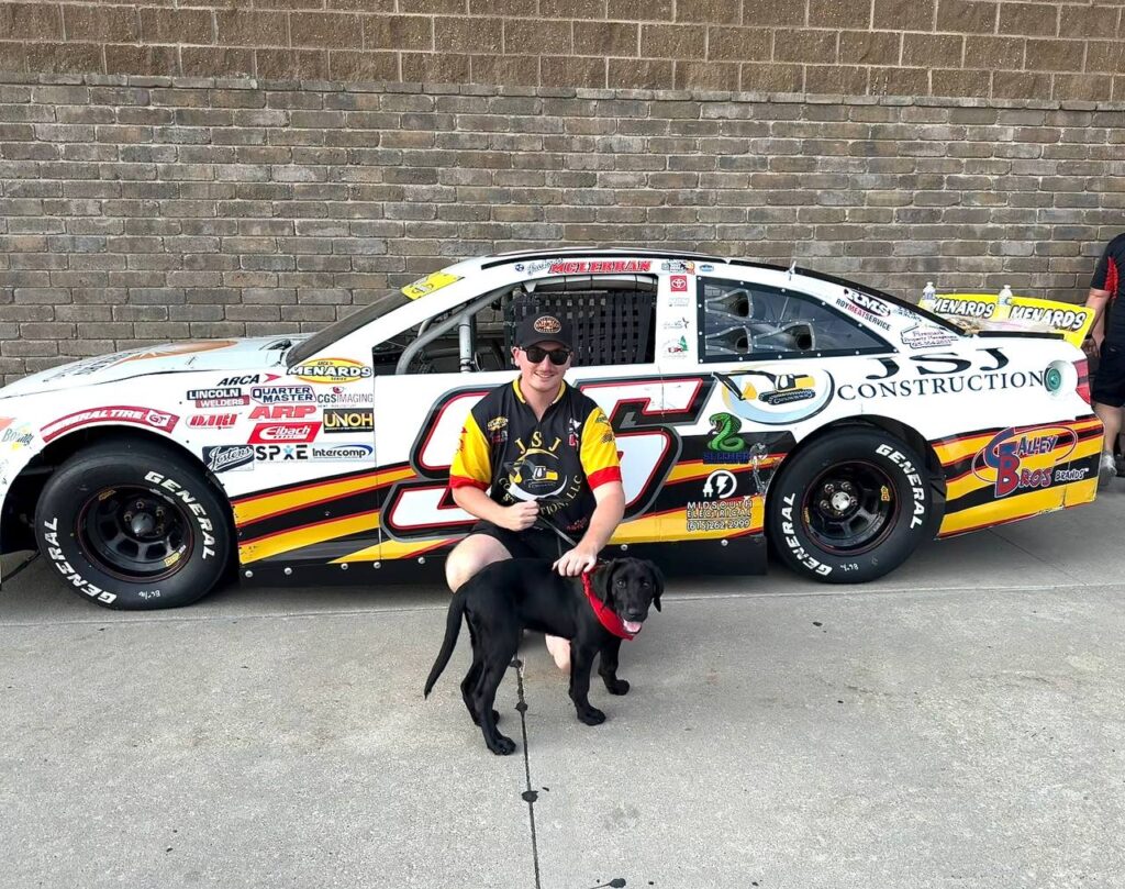 photo_2026-09-14 141551 - Puppy Jake Foundation Man in a black-yellow racing jersey kneels beside a black dog in front of a white, sponsor-laden stock car (number 25).